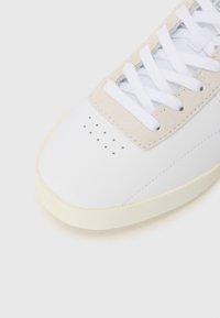 Biele tenisky s hladkým koženým vrchom, svetlosivými semišovými detailmi, bielymi šnúrkami, perforovanou špičkou a krémovou gumovou podrážkou.