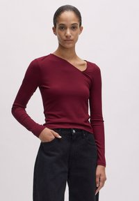 Burgunderfarbener Langarm-Top mit geripptem Material. Verfügt über einen asymmetrischen Ausschnitt und eine figurbetonte Silhouette. Kombiniert mit hoch taillierten schwarzen Jeans.