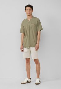Maglietta polo verde a coste con taschino sul petto, maniche corte e scollo a V, abbinata a pantaloni corti color crema e sneakers bianche con accenti neri.