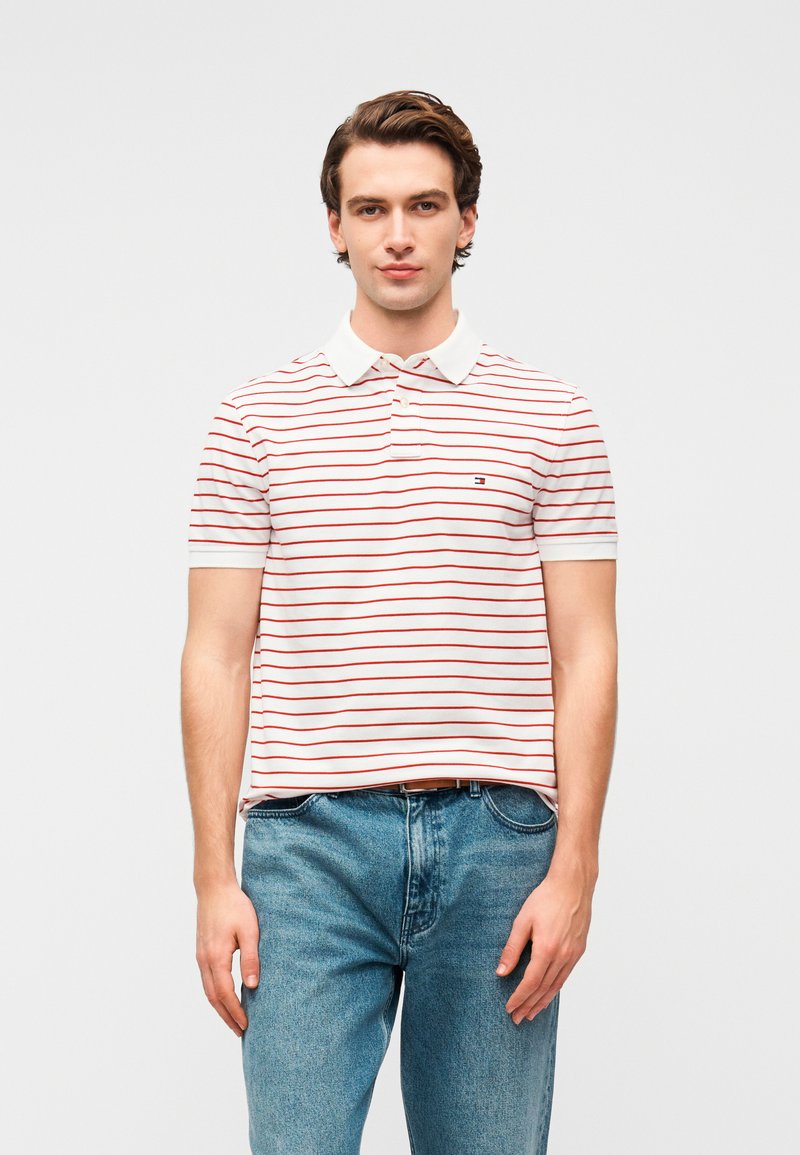 Polo rayé blanc à rayures rouges, doté d'un col et d'un petit logo sur la poitrine gauche. Porté avec un jean denim bleu.