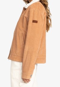 Veste en velours côtelé beige clair avec un col en peau lainée, des poches latérales et un logo Roxy sur la manche gauche. Tissu côtelé texturé.