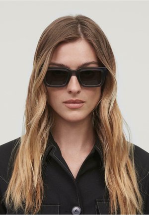 Lunettes de soleil noires rectangulaires avec des montures épaisses. Texture lisse. Portées avec une veste sombre à col boutonné. Cheveux longs et ondulés.