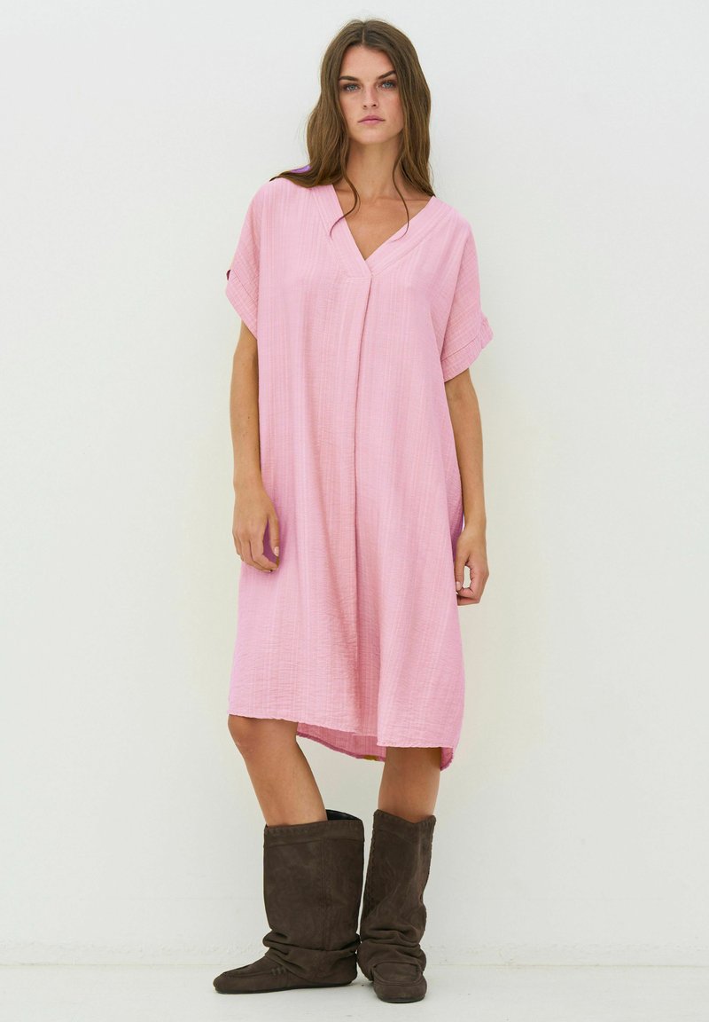 Femme portant une robe ample rose clair à col en V avec manches retroussées et bottes souples brun foncé, debout sur un fond blanc uni.