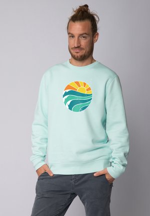 Muntgroene sweatshirt met een cirkelvormige grafiek met een zon en golven in oranje en teal. Zachte textuur met ribgebreide manchetten en zoom.