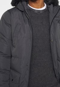 Czarna puchowa kurtka z kapturem, z futrzanym wykończeniem, zamkiem błyskawicznym z przodu i kieszenią na napy. Warstwowa pod teksturowanym swetrem.