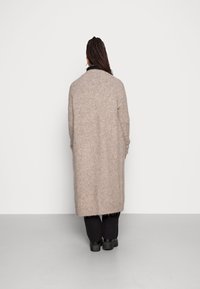 Long gilet surdimensionné en tissu beige doux, finition texturée, devant ouvert avec un ourlet droit, accompagné d'un pantalon noir et de bottes noires épaisses.