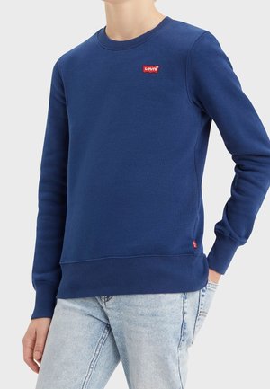 Personne portant un sweat-shirt bleu Levi's avec un patch logo rouge sur la poitrine et un jean bleu clair, debout avec une main dans la poche.