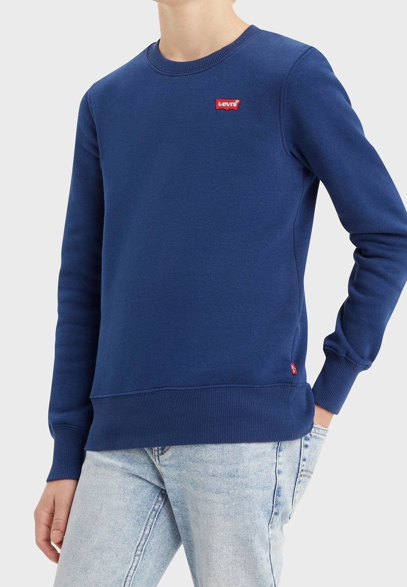 Personne portant un sweat-shirt bleu Levi's avec un patch logo rouge sur la poitrine et un jean bleu clair, debout avec une main dans la poche.
