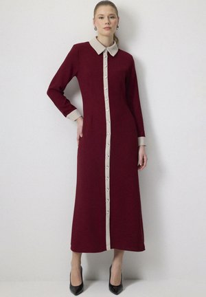 Mujer con un vestido largo burdeos con cuello, puños y tapeta de botones delanteros en beige, posando frente a un fondo claro y liso.
