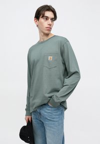 Μακρυμάνικο T-shirt σε απαλό πράσινο βαμβάκι, με τσέπη στο στήθος που φέρει λογότυπο. Συνδυάζεται με ανοιχτόχρωμο τζιν παντελόνι.