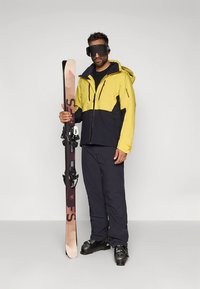 Skiausrüstung mit einer gelb-navyblauen Jacke, schwarzen Hosen und schwarzen Stiefeln. Der Skifahrer hält Skier mit einem pink-beigen Design. Große schwarze Skibrille.