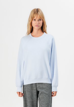 Eine Frau trägt einen hellblauen, langärmeligen Sweatshirt und graue, strukturierte Hose und steht vor einem schlichten weißen Hintergrund.