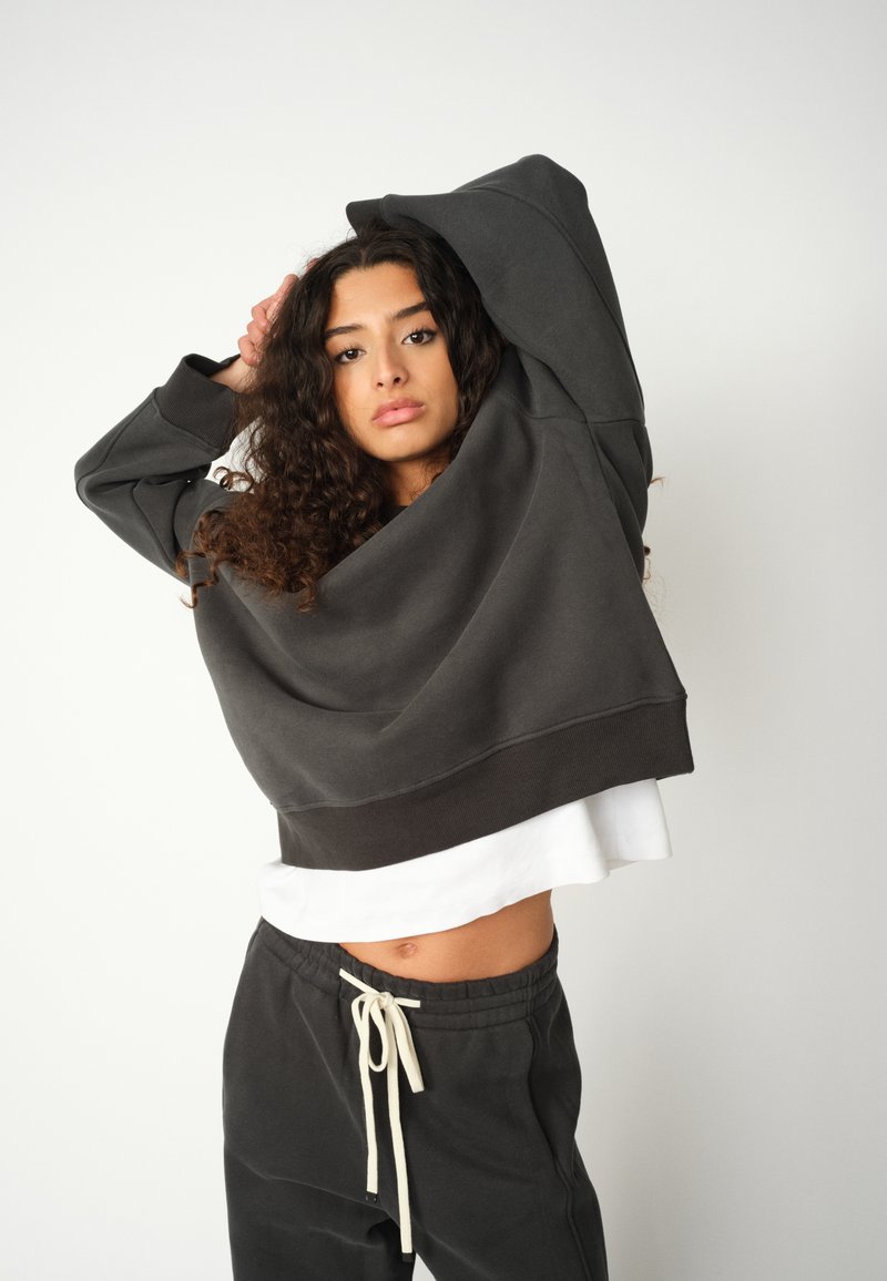 Tmavě šedé zkrácené sweatshirt s širokým výstřihem a žebrovanými manžetami, vrstvené přes bílou košili. V páru s volnými šedými tepláky.