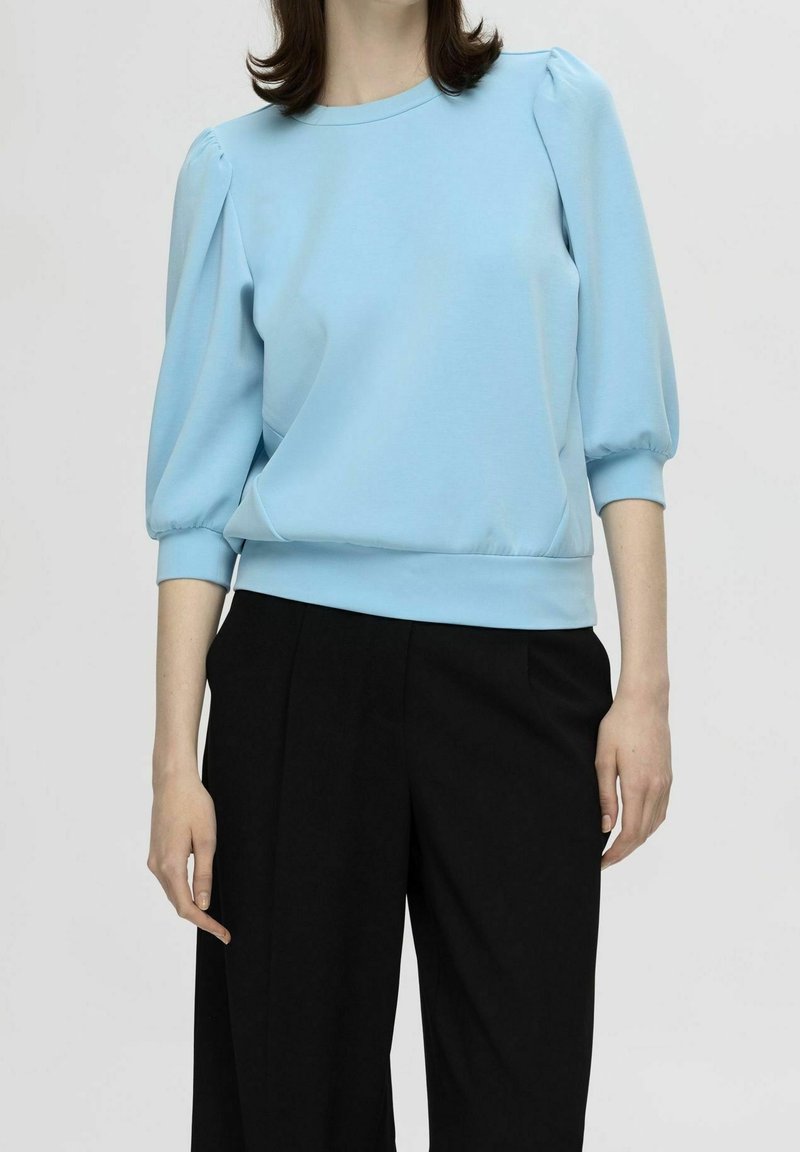Jersey de color azul claro con mangas abullonadas, escote redondo y puños elásticos, combinado con pantalones negros de corte ajustado. Textura de tela suave.