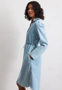 Robe en denim bleu clair avec un col, manches longues, ceinture à la taille et poches latérales, comportant une coupe droite et des coutures discrètes.