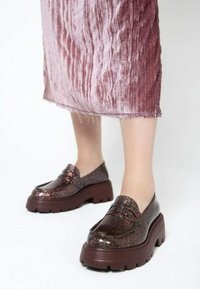 Błyszczące brązowe platformowe mokasyny z grubą podeszwą i gładką fakturą, zestawione ze zmarszczoną różową aksamitną spódnicą.