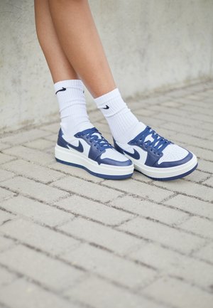 Osoba w białych i granatowych butach sportowych Nike z białymi skarpetkami Nike, stojąca na jasno szarym brukowanym podłożu przy betonowej ścianie.