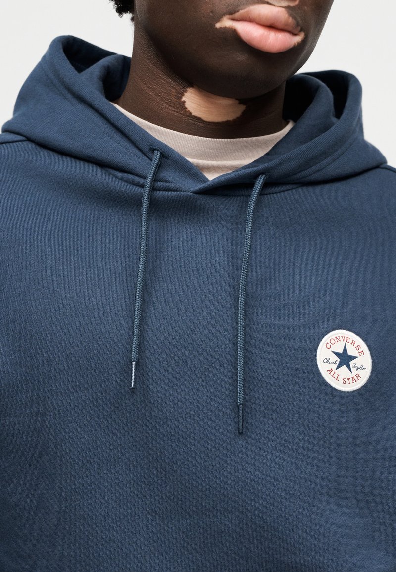 Marineblå hættetrøje med snore, med et rundt "Converse All Star"-mærke. Fremstillet af et tekstureret stof og har en afslappet pasform.