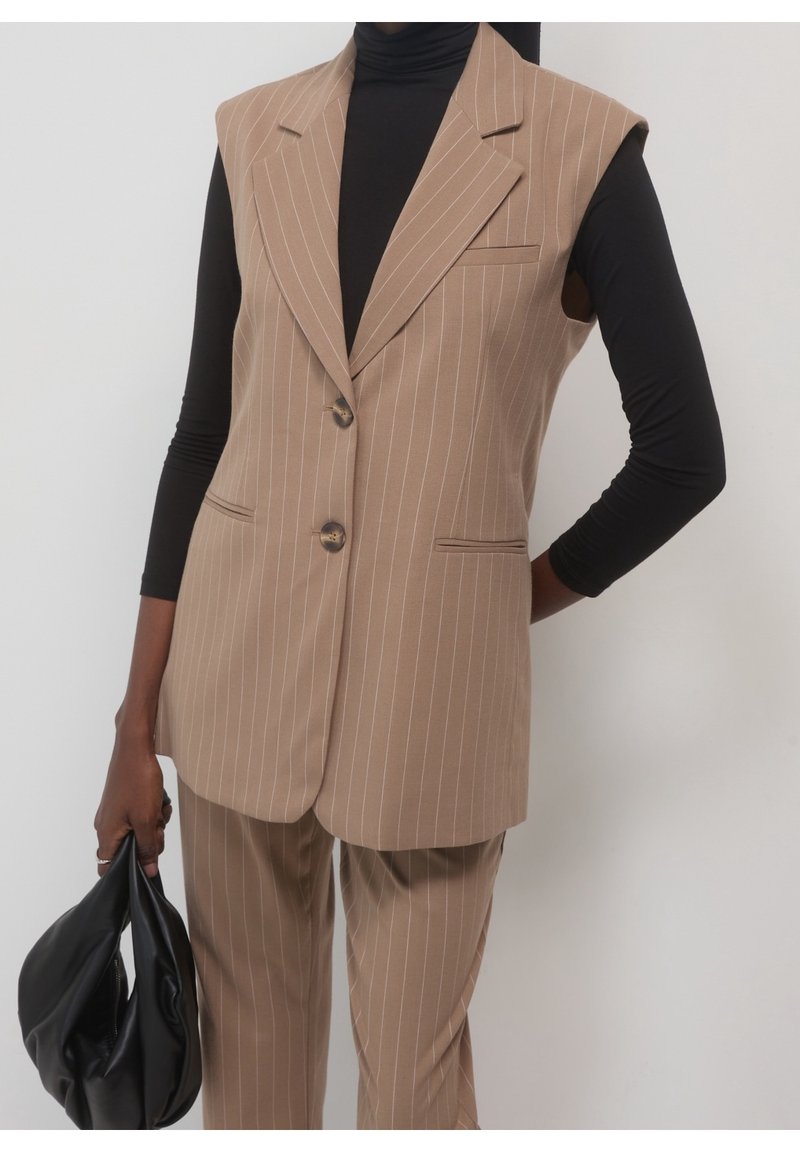Personne portant un blazer sans manches beige à fines rayures et un pantalon assorti sur un col roulé noir, tenant un sac à main en cuir noir dans la main gauche.