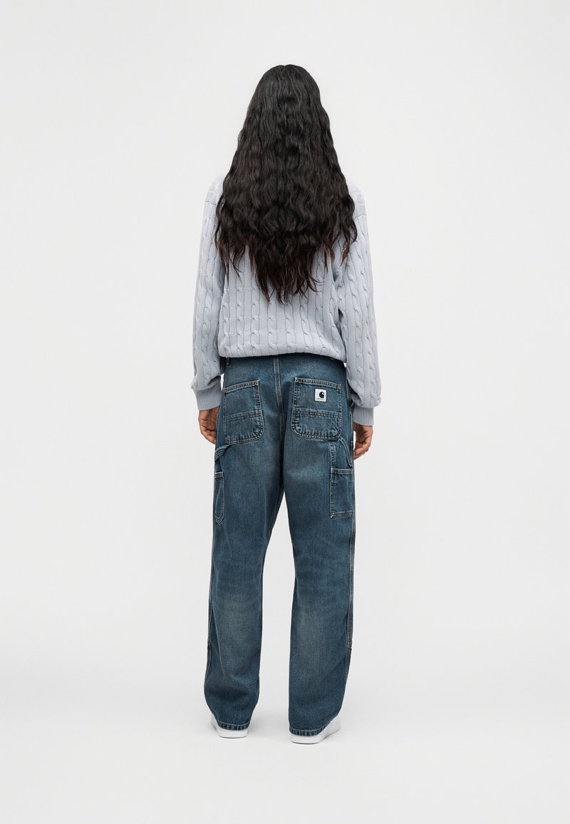 Personne aux cheveux longs et ondulés portant un pull gris clair en maille torsadée et un jean bleu large, debout de dos.