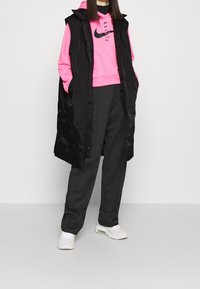 Svart lång väst över en rosa hoodie med en logo, kombinerat med lösa grå byxor och vita sneakers med en subtil orange detalj.