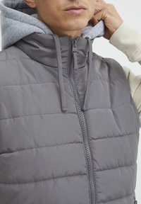 Grå polstret vest med høy krage og frontglidelås. Båret over en lys grå hettegenser, har snorer og en jevn, matt tekstur.