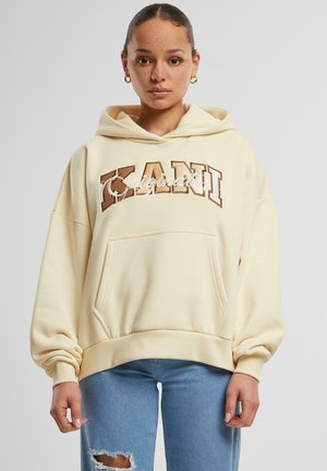Kremfarget hettegenser med frontlomme, oversized ermer, og "KANI Originals" grafikk i brunt og hvitt. Mykt bomullsmateriale.