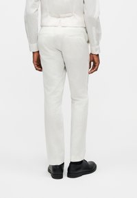 Άνδρας με λευκό παντελόνι φόρμας, λευκό πουκάμισο φόρμας και μαύρα επίσημα παπούτσια, εμφανιζόμενος από πίσω σε φωτεινό φόντο.