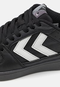 Sorte sneakers med en glatt skinnoverside, teksturerte svarte lisser og hvite chevron-detaljer. Gummisåle med flat profil.