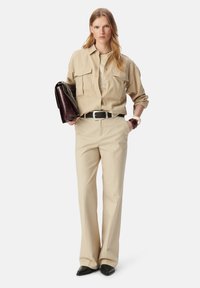 Beige overhemd met knopen en twee borstzakken, beige pinstripe broek, brede zwarte riem en donker handtasje met ketting. Zwarte puntige schoenen.
