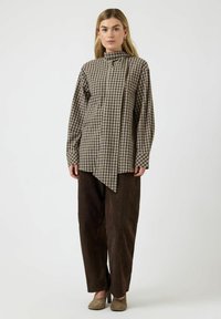 Brun ternet lang skjorte med høj krave og asymmetrisk kant, kombineret med mørkebrune bukser med brede ben og taupefarvede sko.