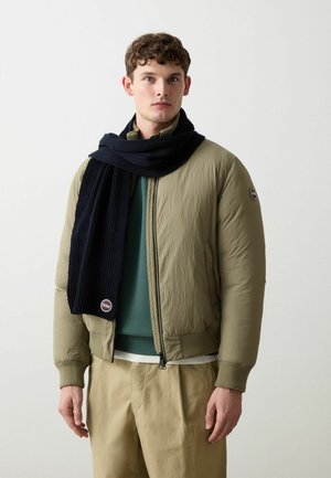 Giacca bomber imbottita verde oliva con polsini a costine, abbinata a una sciarpa lavorata a maglia blu scuro. Design strutturato, chiusura con zip e patch del logo sulla manica.