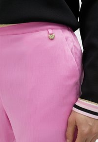 Rosa byxor med en lätt krinklad textur, med en dekorativ knapp i guldton formad som ett hjärta på midjan och svart tyg i närheten.