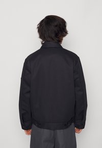 Svart jacka i slätt tyg, med klassisk krage, knappstängning vid midjan och sidofickor, sedd från baksidan.