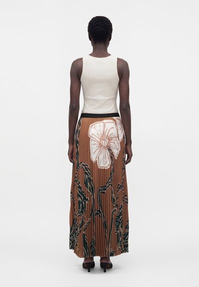 Femme aux cheveux courts portant un haut blanc sans manches et une jupe longue plissée marron avec de grands imprimés floraux blancs, debout, de dos.