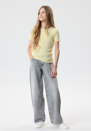 Jeune personne aux cheveux longs portant un t-shirt jaune pâle, un jean gris à jambes larges avec de petits points blancs, et des baskets blanches, debout de manière détendue.