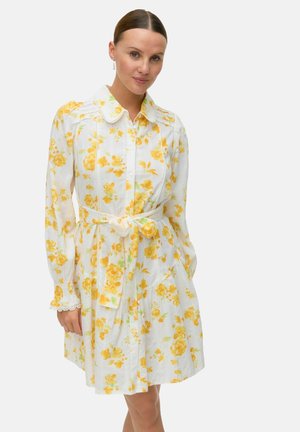 Robe blanche avec imprimé floral jaune, manches longues et col. Présente une taille à nouer et de délicates accents en dentelle sur les manches.