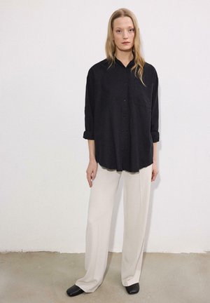 Femeie cu păr lung și blond, purtând o cămașă neagră largă cu nasturi, pantaloni bej largi și pantofi negri, stând în fața unui perete alb simplu.