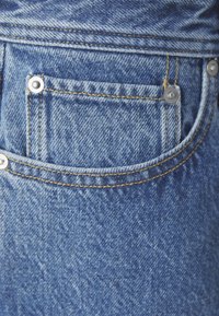 Blå denimtyg med en bakficka med silvernitar och gul söm. Texturen är något urblekt med en mjuk finish.