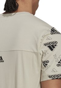 Vaalea beige, lyhythihainen paita, jossa on mustat Adidas-logot ja abstrakteja kuvioita hihoissa, sileä rakenne ja säännöllinen istuvuus.