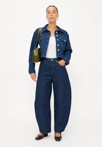 Denimjack met knoopsluiting, ruwe zoom, gecombineerd met brede, donkerblauwe denim jeans. Een groene schoudertas en zwarte ballerina's maken de look compleet.