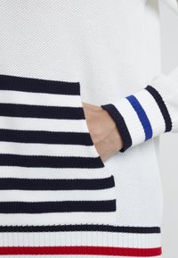 Vit stickad tröja med marinblå och vita randiga detaljer, med en randig ficka och ribbade mudd med röda och blå ränder.