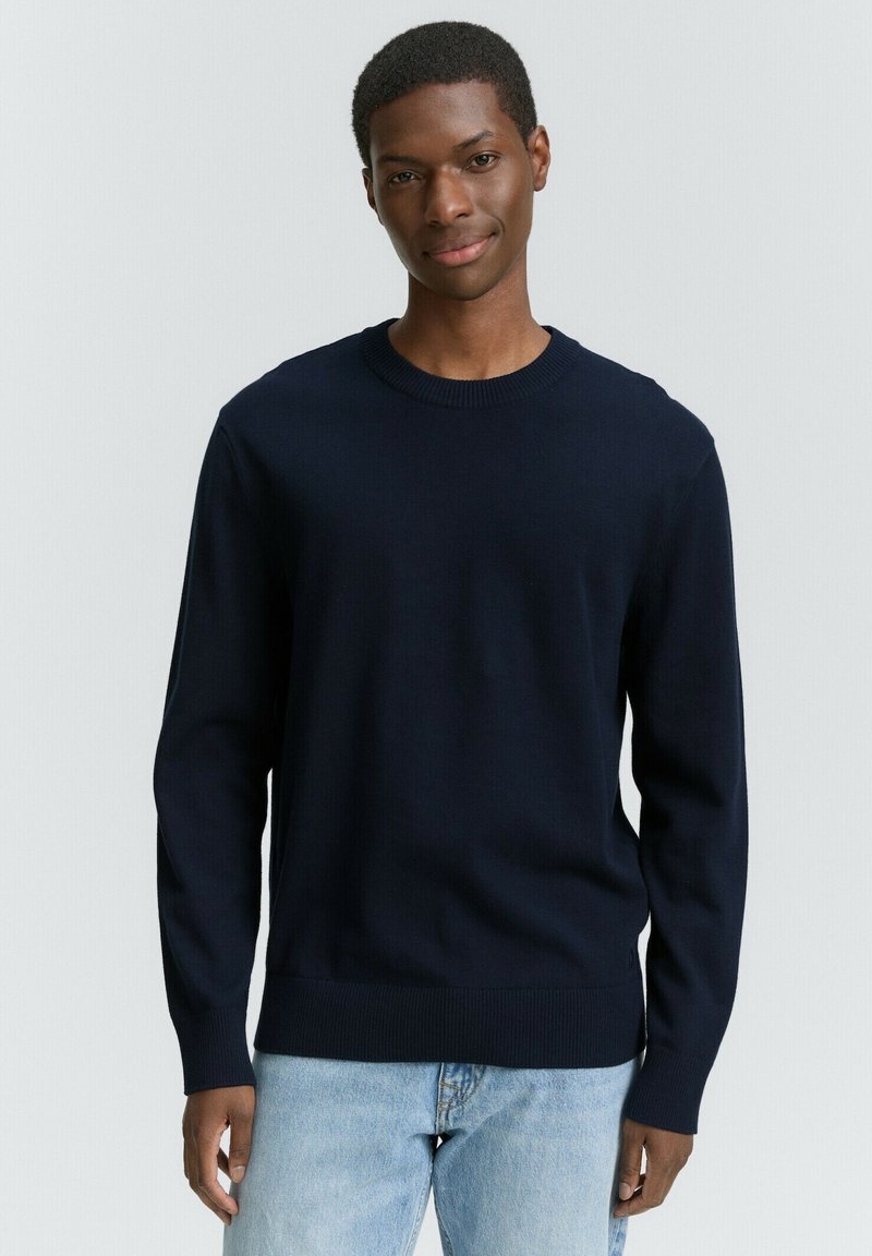 Jonge man in een marineblauwe trui en lichtblauwe jeans, glimlachend tegen een eenvoudige lichte achtergrond.