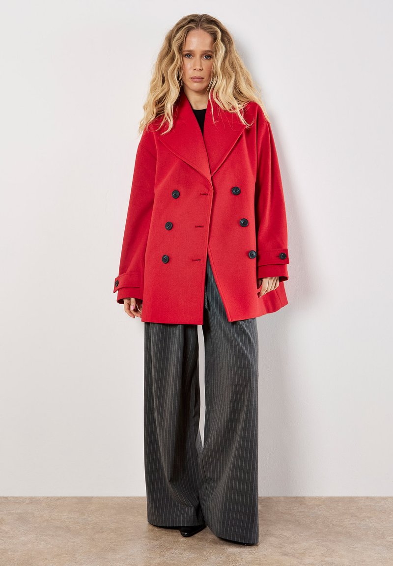 Punainen kaksirivinen takki, jossa on ylimitoitetut kaulukset, mustat napit ja rento siluetti, yhdistettynä leveälahkeisiin raitapicturetakkahousuihin.