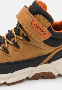 Chaussure de randonnée marron et noire avec une cheville rembourrée, doublure en maille, semelle en caoutchouc texturée et sangle ajustable avec une étiquette orange vif.