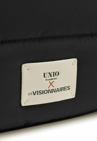 Borsa di stoffa nera con un'etichetta rettangolare crema con testo in rilievo "UNIO HAMBURG X LES VISIONNAIRES" e cuciture di fissaggio.