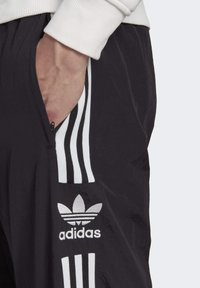 Mustat urheiluhousut, joissa on kolme valkoista raitaa sivuilla, varustettuna vetoketjullisella taskulla ja metallinhohtoisella hopean värisellä adidas-logolla edessä.