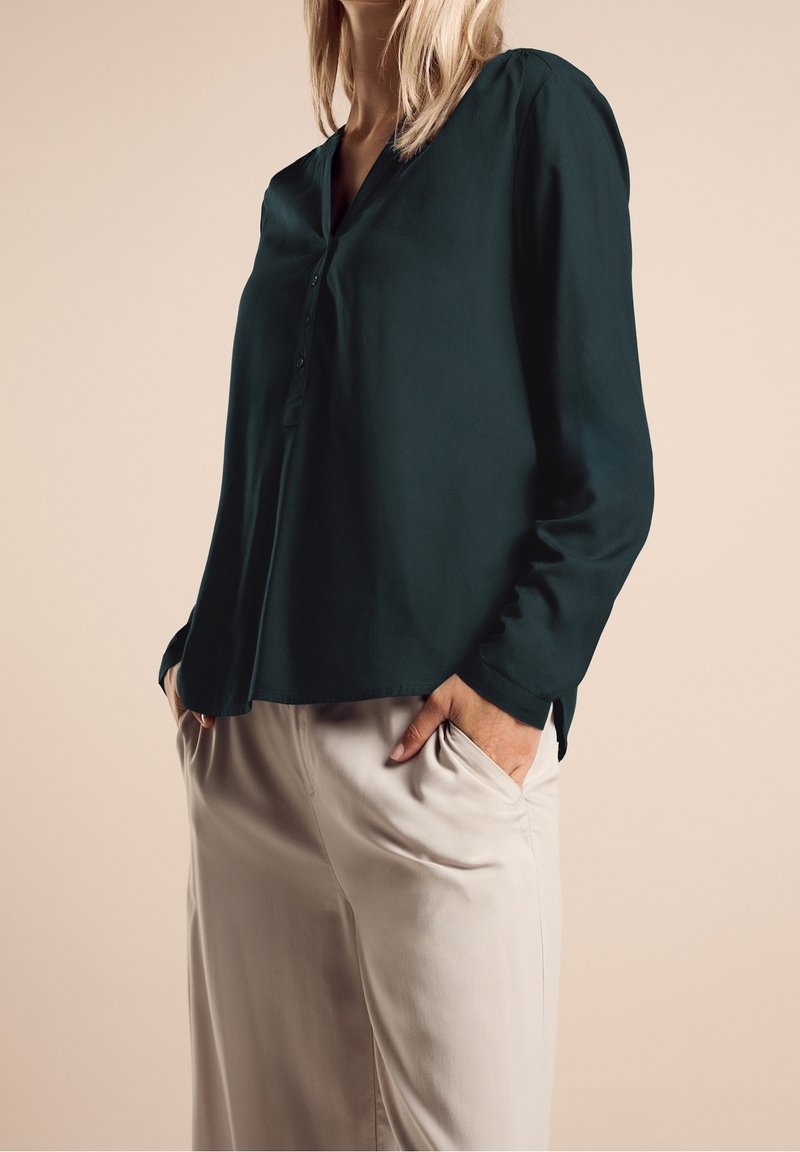 Femme portant un chemisier boutonné manches longues vert foncé et un pantalon ample beige clair, les mains dans les poches, sur fond beige.