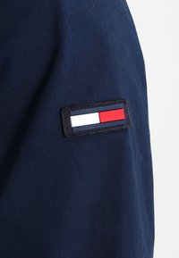 Marinblått tyg med en liten rektangulär lappar som har röda, vita och blå ränder. Lappen är sydd på överarmen av plagget.