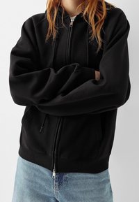 Sudadera con cremallera negra de textura suave, con puños y dobladillo acanalados, y bolsillos a los lados, combinada con jeans de un azul claro.